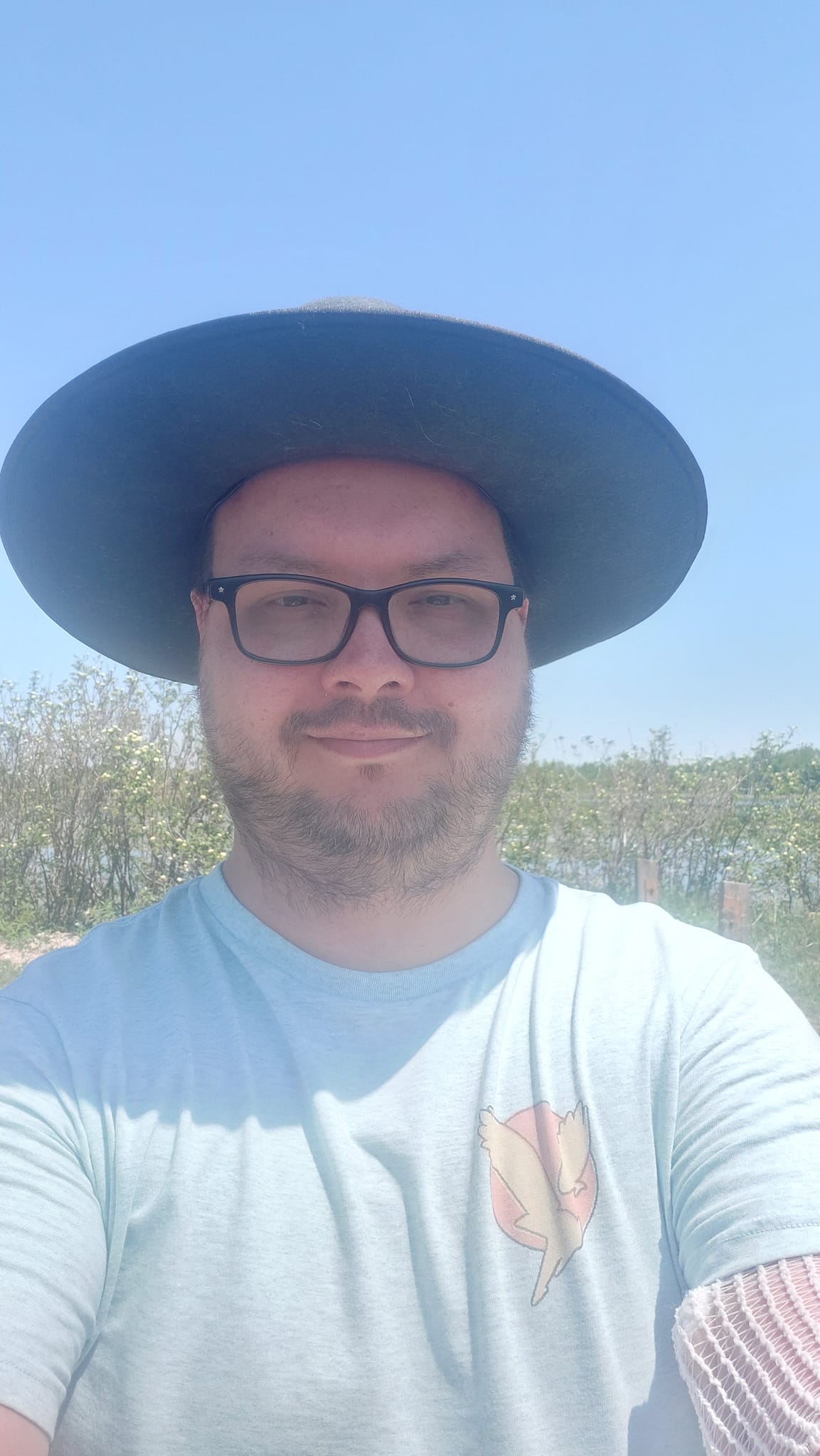 A man wearing a blue shirt wears a wide brimmed sunhat in front of a wetland